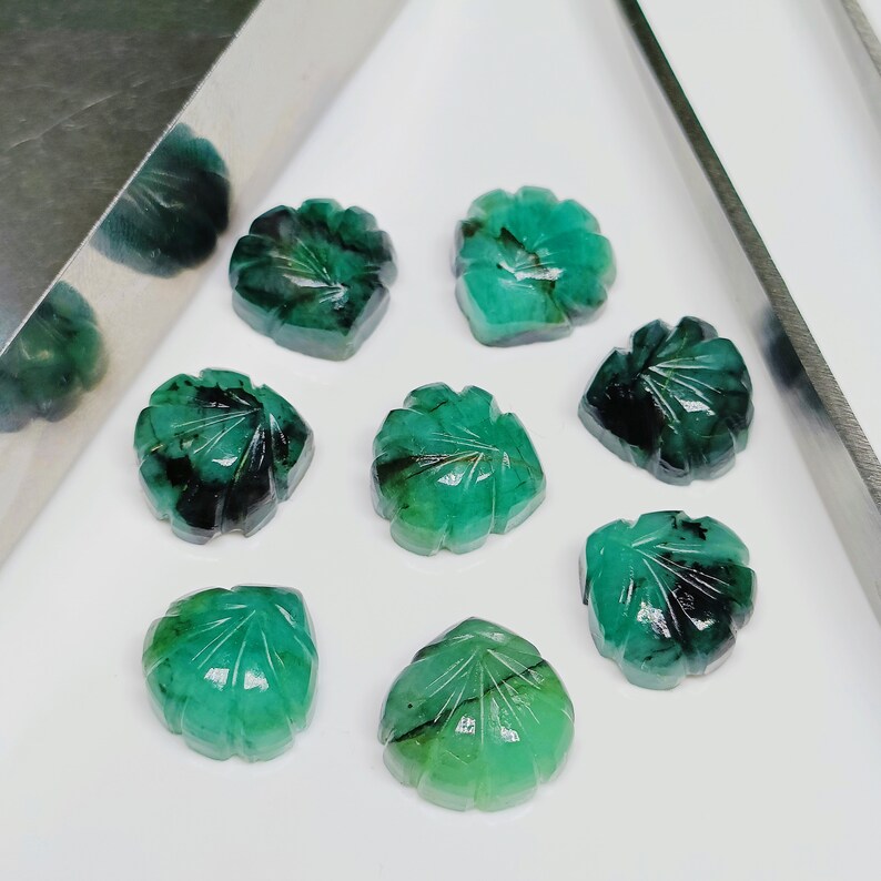 Puede incluir: Ocho piedras preciosas de esmeralda verde en forma de hoja con inclusiones negras. Las piedras preciosas est&aacute;n dispuestas en semic&iacute;rculo sobre una superficie blanca.