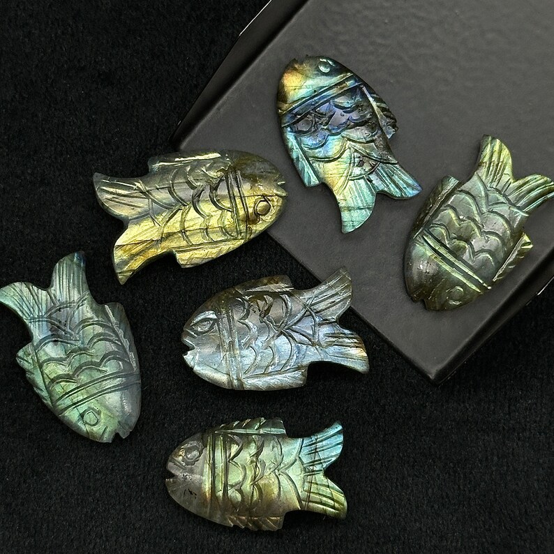Puede incluir: Seis tallas de peces de labradorita iridiscente, cada una con un dise&ntilde;o &uacute;nico y detalles intrincados. Las tallas se muestran sobre una superficie negra.