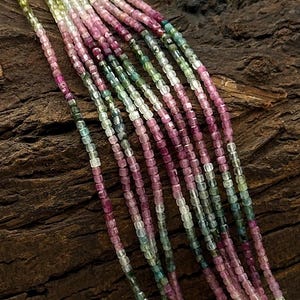 Puede incluir: Una hebra de cuentas de piedras preciosas multicolores, con tonos de rosa, verde y blanco. Las cuentas son de forma cuadrada y tienen un acabado liso y pulido.