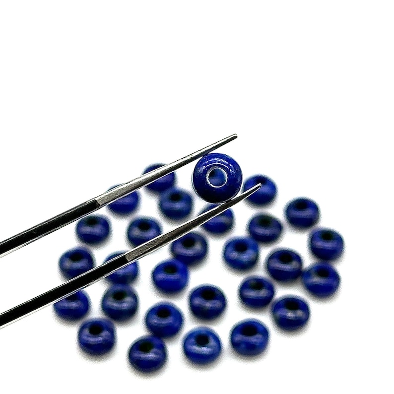 May include: A close-up of a single blue gemstone bead held by tweezers, with a scattering of similar beads in the background.