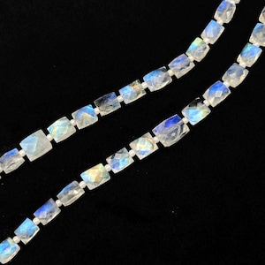 Op de afbeelding: Twee strengen rechthoekige, iriserende maansteen kralen tegen een zwarte achtergrond. De kralen zijn een mix van wit, helder en blauw, met een facetgeslepen snit die het licht vangt. De kralen zijn geregen op een witte draad.