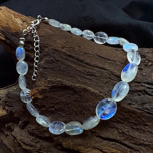 Puede incluir: Un collar de cadena de plata con cuentas de piedra lunar de forma ovalada. Las cuentas son translúcidas y tienen un brillo iridiscente azul.