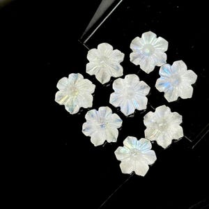 May include: A collection of eight iridescent, flower-shaped beads. Each bead has a delicate, translucent appearance with subtle color shifts. The beads are arranged on a black surface, creating a contrast that highlights their intricate details.