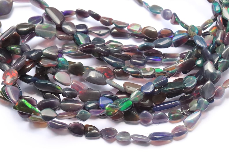 May include: A strand of iridescent black opal gemstone beads. The beads are irregularly shaped and have a smooth, polished finish. The beads are strung on a clear thread and are ready to be used for jewelry making.