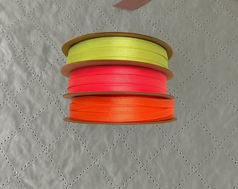 May include: Three spools of brightly colored ribbon in neon yellow, hot pink, and orange. The ribbon is stacked on top of each other, with the spools made of cardboard. The background is a quilted gray fabric.