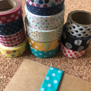 May include: A stack of colourful washi tapes with polka dot patterns, including red, blue, yellow, gold, and black. The tapes are arranged in a vertical stack on a cork surface. A teal blue washi tape with white polka dots is lying on a brown paper surface.