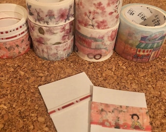 May include: A collection of decorative washi tapes with floral and illustrated designs. The tapes are rolled and stacked on a cork surface. There are two blank cards with washi tape samples on them.