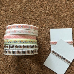 May include: A stack of five rolls of decorative washi tape with floral and geometric patterns. The tape is on a cork background. Two white cards are also shown with a strip of washi tape on each.