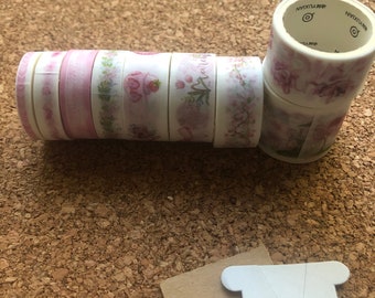 May include: A stack of eight rolls of decorative washi tape with floral patterns in pink and green. The tape is wrapped around a cardboard spool. The tape is on a cork surface.