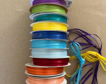 May include: A stack of spools of ribbon in various colors, including white, black, purple, green, turquoise, yellow, blue, light blue, white, red, orange, peach, pink, and light pink. The spools are stacked on top of each other, and there are two loose pieces of ribbon in the bottom right corner of the image.