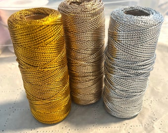 May include: Three spools of metallic thread in gold, silver, and a light gold color. The spools are sitting on a white surface.