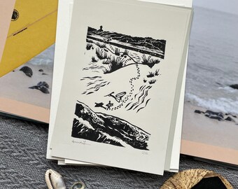 May include: Black and white linocut print of a beach scene with two figures walking on a path. The print is titled "Seaward" and numbered 2/12.