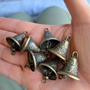 Op de afbeelding: Een verzameling van acht kleine, antiek-gouden bellen met ingewikkelde ontwerpen. De bellen hebben een licht aangetast oppervlak en zijn aan elkaar geregen met een ketting.