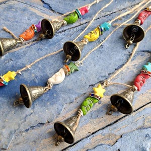 Peut inclure: Carillons décoratifs avec de petites cloches en bronze et des accents de perles colorées. Les perles ont la forme d'étoiles, de croissants de lune et d'autres formes organiques dans des tons de rouge, jaune, vert et turquoise. Les carillons sont suspendus à de la ficelle naturelle.