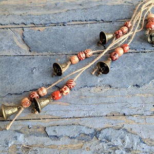 Pode incluir: Uma corda de pequenos sinos de latão com contas de madeira em tons de marrom e vermelho. Os sinos são presos a um cordão de juta natural.