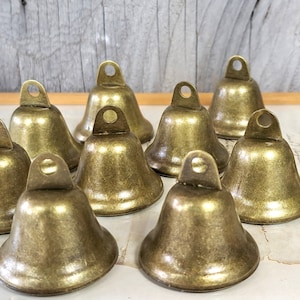 Könnte beinhalten: Eine Gruppe von zehn kleinen, antiken goldfarbenen Glocken mit abgerundeter Form und einer kleinen Schlaufe oben. Die Glocken sind in einer Gruppe auf einer weißen Oberfläche angeordnet.