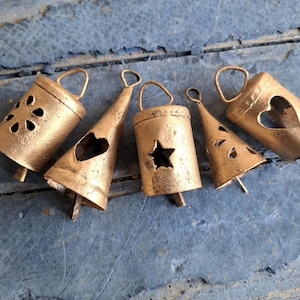 Op de afbeelding: Zes gouden metalen bellen met verschillende uitgesneden ontwerpen, waaronder een hart, een ster en een bloem. De bellen hangen aan een houten oppervlak.