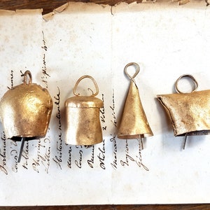 Op de afbeelding: Vijf goudkleurige metalen bellen in verschillende vormen en maten. De bellen hangen aan een stuk papier met handgeschreven tekst.