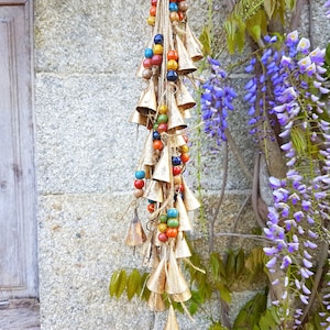 Op de afbeelding: Een reeks gouden bellen met gekleurde kralen die aan een muur hangen. De bellen zijn in een cascade-patroon gerangschikt, met de grootste bellen bovenaan en de kleinste bellen onderaan. De kralen zijn in verschillende kleuren, waaronder rood, blauw, groen en geel.