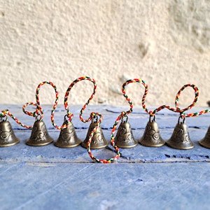 Op de afbeelding: Een set van acht kleine, messingkleurige bellen met ingewikkelde ontwerpen, verbonden door een rood, groen en geel touwtje.