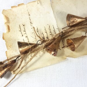 Peut inclure: Une guirlande rustique de petites cloches en bronze vieilli, enfilées sur de la ficelle, posée sur un morceau de papier ancien avec une écriture délavée. Les cloches sont coniques et légèrement ternies. Fond blanc texturé.
