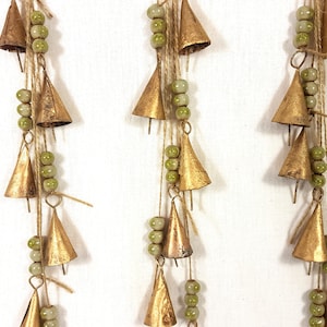 Op de afbeelding: Drie strengen gouden bellen met groene kralen die aan een witte achtergrond hangen. Elke streng heeft een ander aantal bellen en kralen.