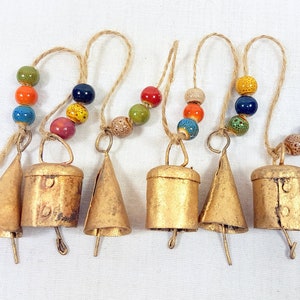 May include: Six gold-coloured bells of varying shapes and sizes hang from twine strings with colourful beads. The bells have a rustic, aged finish.