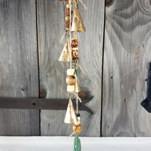 May include: A decorative wind chime with small, gold-coloured bells and wooden beads strung on twine. The chime includes a teal-coloured feather pendant at the bottom. The background is a weathered wooden door.