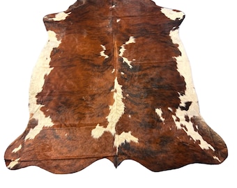 May include: A full view of a cowhide rug. The rug has a natural pattern of brown, white, and black markings. The hide is irregularly shaped, with a soft, textured appearance. The rug is suitable for use as a floor covering or decorative accent.