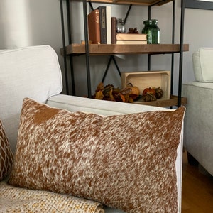 May include: A brown and white cowhide throw pillow with a white background and brown spots. The pillow is on a white couch with a tan throw blanket.