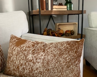 Könnte beinhalten: Ein braunes und weißes Kuhfell-Wurfkissen mit weißem Hintergrund und braunen Flecken. Das Kissen liegt auf einem weißen Sofa mit einer braunen Wolldecke.
