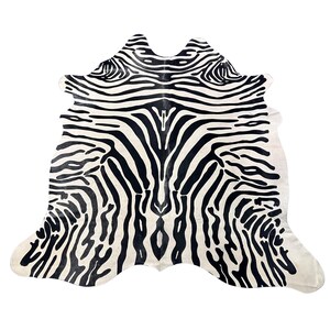 May include: A natural cowhide rug featuring a bold, black and white zebra-inspired striped pattern. The hide has an irregular, organic shape with a symmetrical design radiating from the center, showcasing high-contrast monochromatic lines.