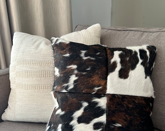 May include: Two throw pillows on a gray couch. One pillow is white with a textured weave. The other pillow is made of brown and black cowhide with a patchwork design.