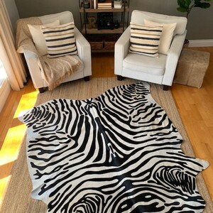 May include: Black and white zebra print cowhide rug with a contemporary design. The rug is laid out on a wooden floor in a living room setting with two white armchairs and a wooden shelf unit.