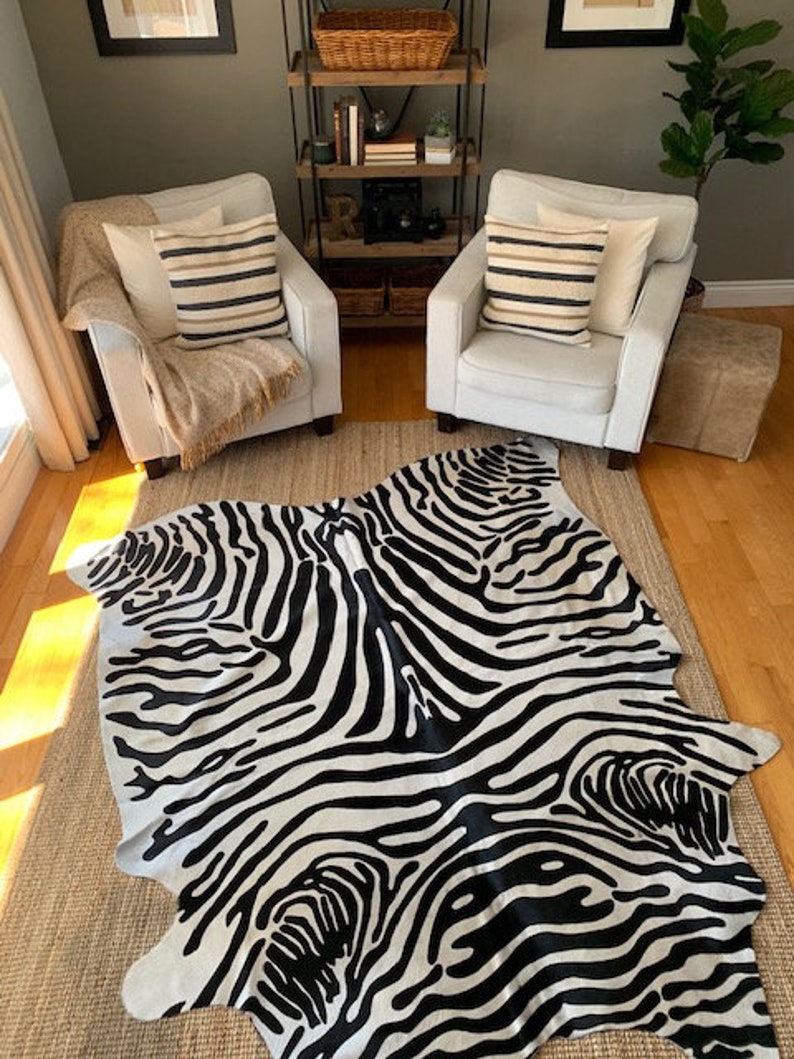 May include: Black and white zebra print cowhide rug with a contemporary design. The rug is laid out on a wooden floor in a living room setting with two white armchairs and a wooden shelf unit.