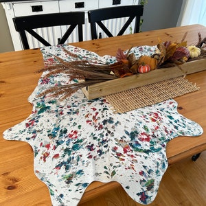 May include: A white cowhide rug with colorful splatter paint design, used as a table runner on a wooden table with a wooden box filled with fall foliage and pumpkins.