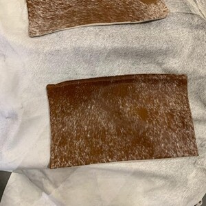May include: Two brown and white cowhide rectangular pillows. The pillows are on a white surface.