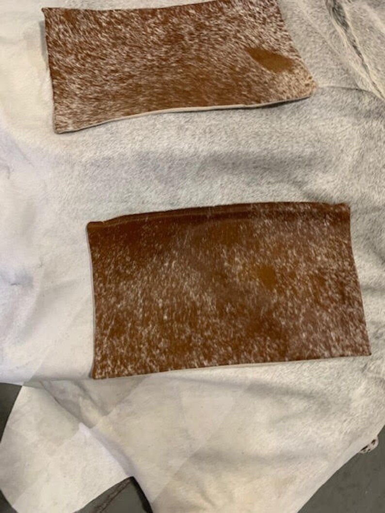 May include: Two brown and white cowhide rectangular pillows. The pillows are on a white surface.