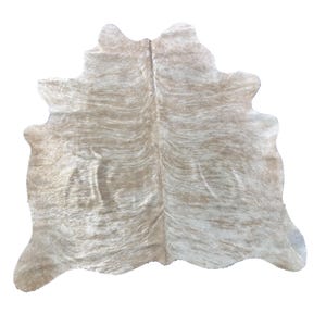 May include: A cowhide rug in beige and white, with a natural, uneven shape. The rug displays a pattern of light and dark brown streaks, giving it a textured look. The hide is spread out, highlighting its full shape and colour variations.