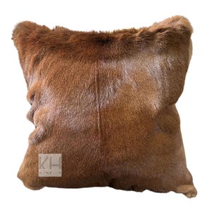 May include: A square, brown fur pillow with a soft, textured appearance. The pillow has a natural, earthy tone and is likely made from animal hide. The pillow is a decorative item for home decor.