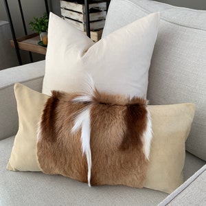 May include: A brown and white fur throw pillow with a beige suede backing on a light gray couch.