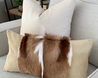 May include: A brown and white fur throw pillow with a beige suede backing on a light gray couch.