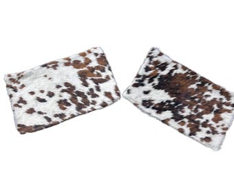 May include: Two rectangular cowhide pillows. The pillows have a white background with brown spots and patches. The pillows are fluffy and soft, perfect for a rustic or farmhouse-style home decor.