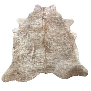 May include: A natural cowhide rug in light brown and cream hues. The rug has an organic shape with rounded edges and a textured surface. This cowhide is a decorative floor covering, suitable for any room.