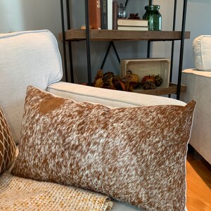 May include: A brown and white cowhide throw pillow with a soft, plush texture. The pillow is sitting on a white couch with a tan throw blanket.