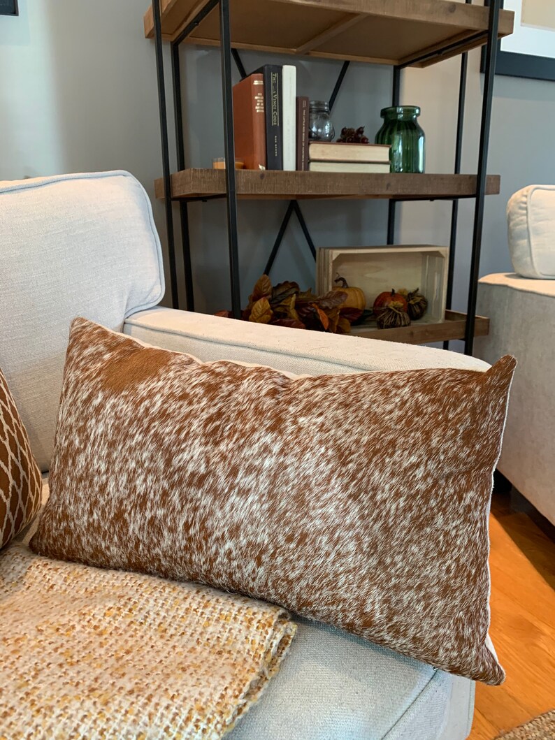 May include: A brown and white cowhide throw pillow with a soft, plush texture. The pillow is sitting on a white couch with a tan throw blanket.