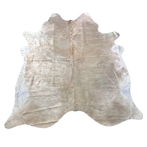May include: A cowhide rug in natural shades of beige, tan, and white. The rug has an organic shape, with a soft, textured feel. The hide is likely from a cow, and the pattern is unique to each piece.