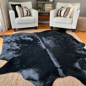 May include: A black cowhide rug with a natural, shaggy texture. The rug is laid out on a woven rug with a light brown color. Two white armchairs with black and white patterned throw pillows are placed on either side of the rug.