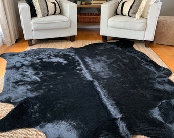 May include: A black cowhide rug with a natural, shaggy texture. The rug is laid out on a woven rug with a light brown color. Two white armchairs with black and white patterned throw pillows are placed on either side of the rug.
