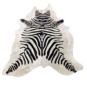 May include: A hide rug featuring a white base with a bold, symmetrical black zebra stripe pattern. The rug has an irregular, organic shape with curved edges, showcasing a high-contrast design that radiates from the center outward.